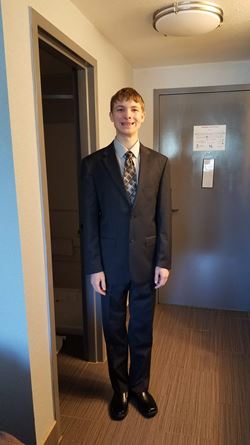 Ryan wearing a suit., standing in a hallway. He is smiling.